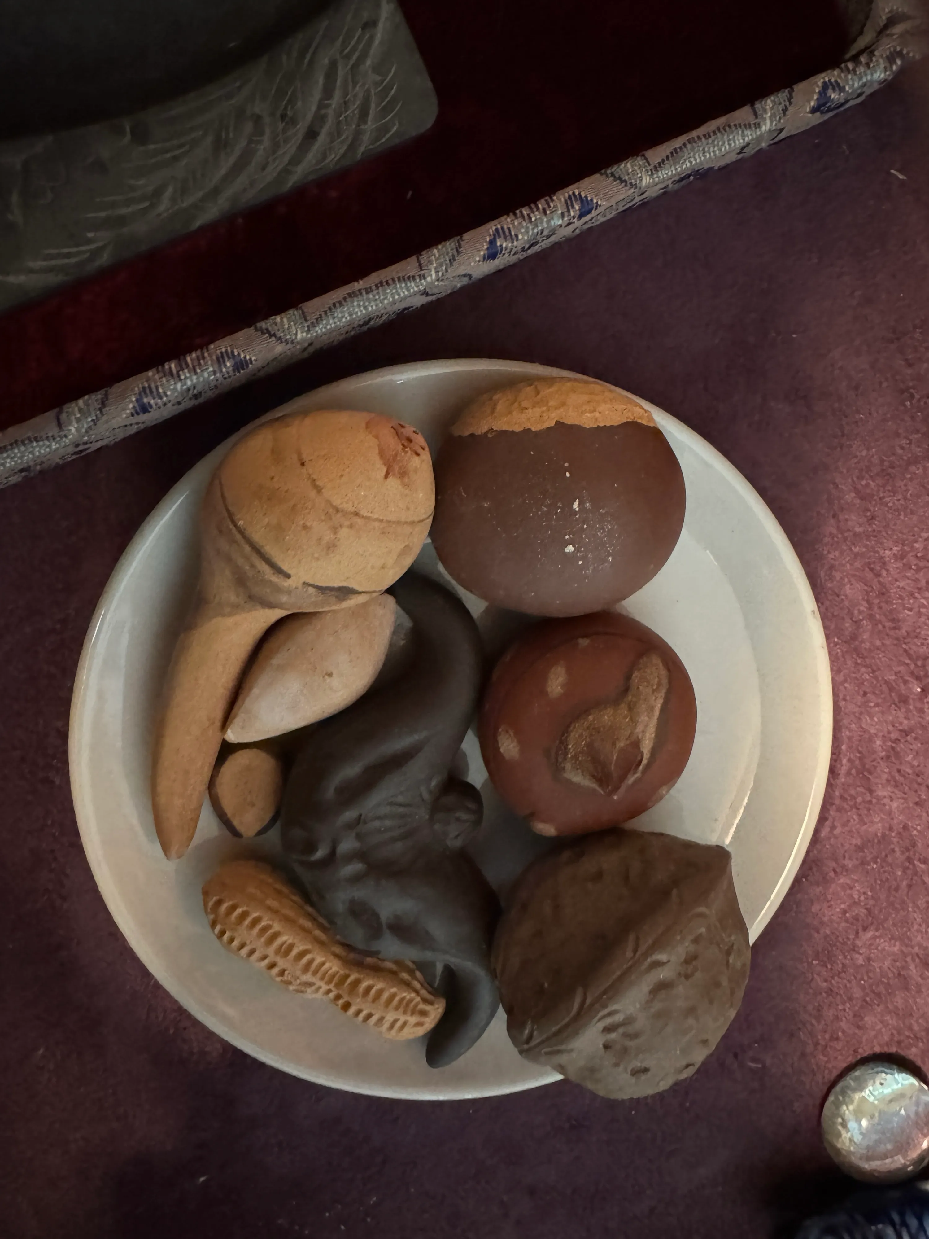 Six small unglazed clay sculptures shaped like nuts, seeds, and a curled mouse, arranged together on a pale ceramic dish.